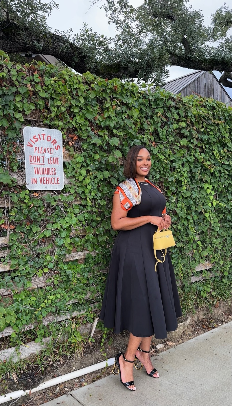 Vanessa in a black dress with a yellow bag in front of an ivy wall